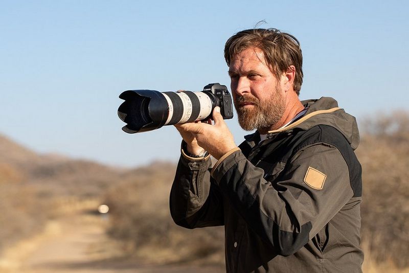 Photographer Brent Stirton stands in a Namibian landscape holding a Canon EOS R with a 70-200mm lens and lens hood attached.