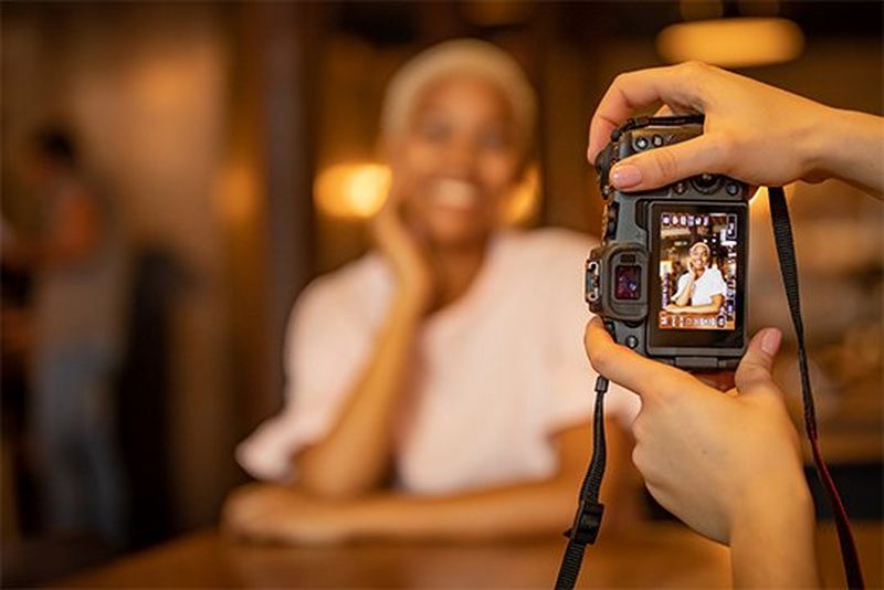 A photographer&#39;s hands hold a Canon EOS RP as she photographs a smiling woman in a dimly-lit interior.