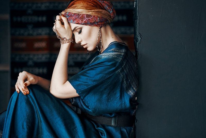 A woman in a dark blue silk dress and a patterned headscarf sits with her back against a wall and her forehead resting in her hand. Taken by Jaroslav Monchak.
