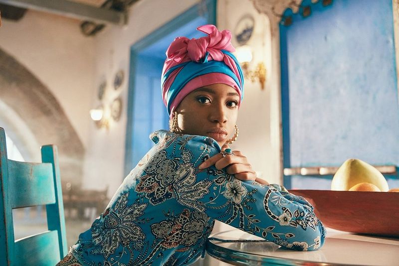 A woman in a blue and pink headscarf sits at a table, resting her elbows on the table with her arms crossed. Taken by Jaroslav Monchak.