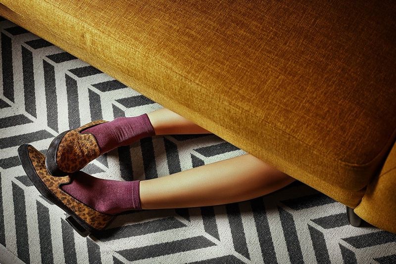 A pair of feet, in purple socks and animal-print loafers, poking out from underneath a mustard velvet sofa. Taken by Jaroslav Monchak.