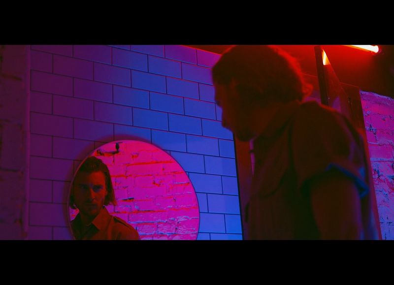 A man in a room lit by blue and magenta neon light stares at his reflection in a circular mirror on the tiled wall. Taken on a Canon EOS R by Jaroslav Monchak.