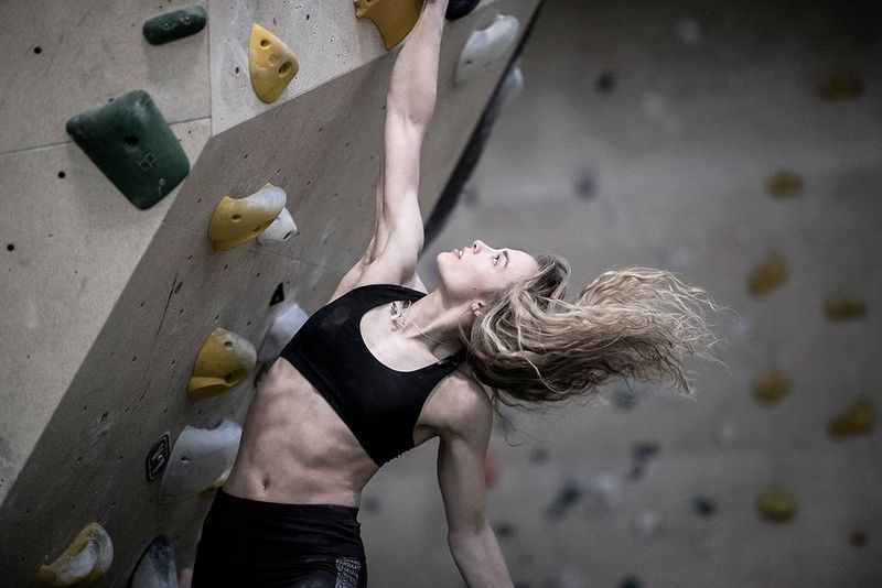 Climber Imogen Horrocks hangs from a climbing wall by one hand. Photo by Julian Finney.