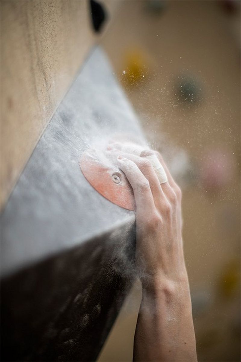 A close-up of an athlete's hand, powder-covered fingertips clinging to a hold in a climbing wall.