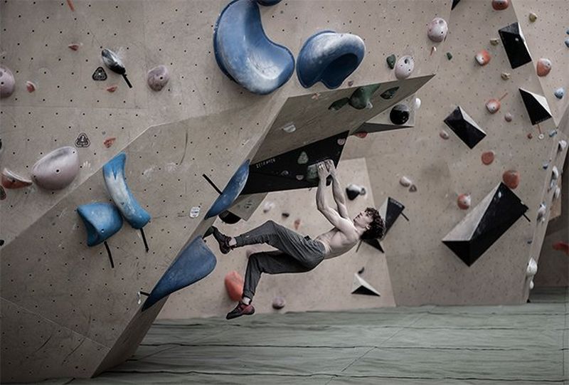 A male athlete hangs almost horizontal under a climbing wall, one leg in the air.