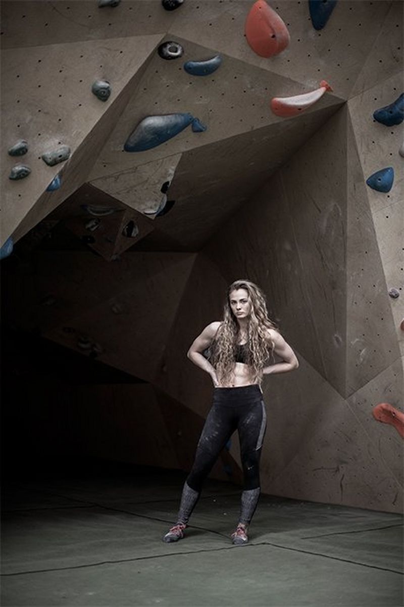 A female climber stands, hands on hips, under a climbing wall with colourful handholds.