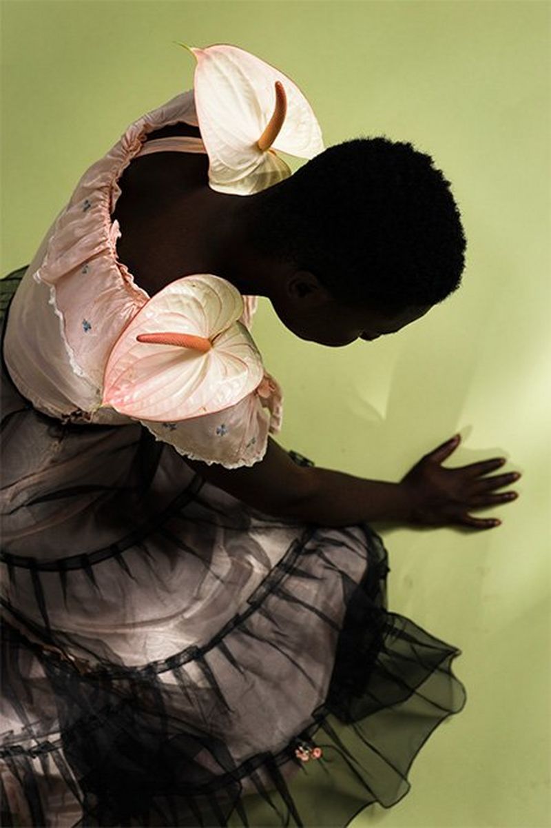 A black model wearing a pink dress with a black overlay leans forward against a light green background with flowers placed on each of her shoulders. 