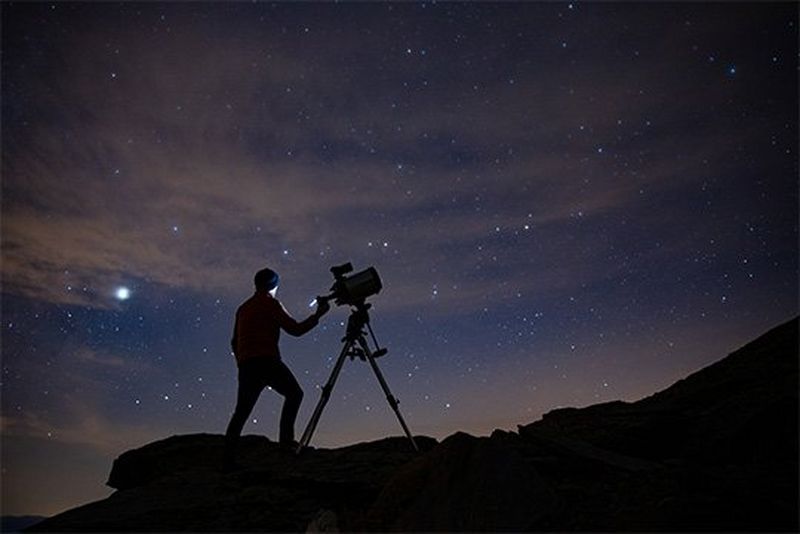 Silhouetted against the night sky, an astrophotographer stands on steep terrain with a Canon EOS Ra attached to a telescope.