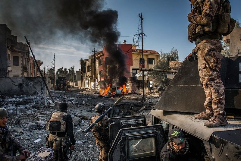 Soldiers stand on and around a tank in a bombed-out city street; a car on fire a few metres away.