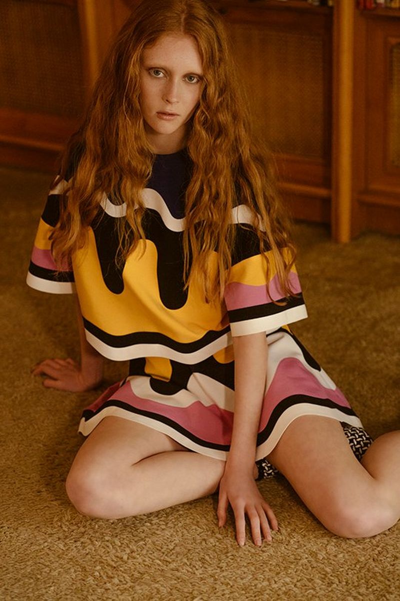 A women sits down on a carpet, looking directly into the camera, her left hand resting on the carpet in front of her.