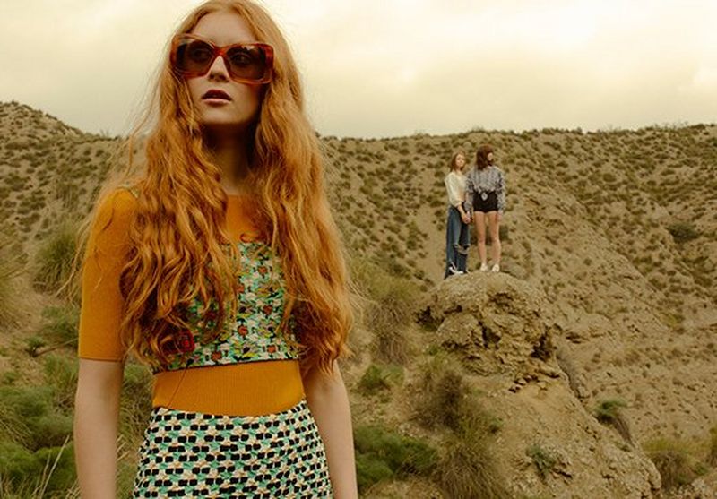 A female model stands outside in the foreground, with two other female models standing on a hill in the background.