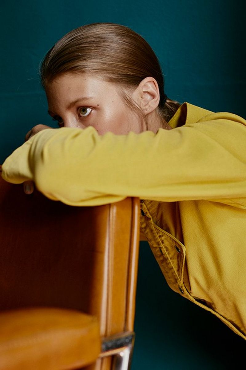 Resting her head on her left arm, balanced on the back of a light tan leather chair, a woman stares ahead.
