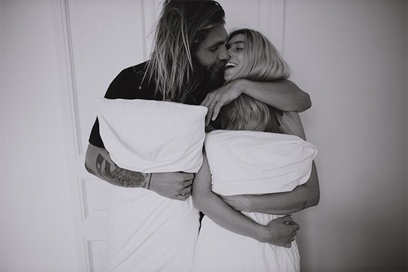 The couple stand side-by-side holding pillows to their stomachs, laughing.