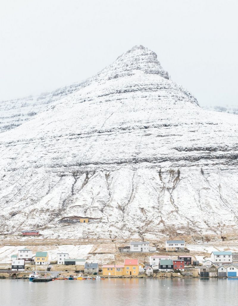 Brightly painted wooden buildings on the waterfront are dwarfed by a huge, snow-covered mountain rising steeply from the shore.