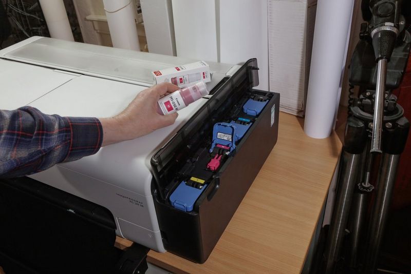 Een hand met daarin een fles pigmentinkt in de buurt van de open kleurentank van een grootformaat printer van Canon.