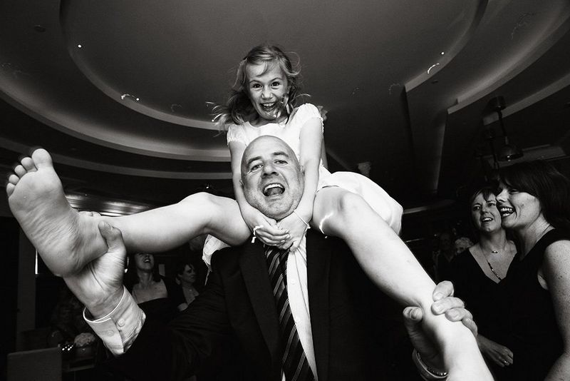 A man dances with his daughter on his shoulders, both laughing and looking at the camera.
