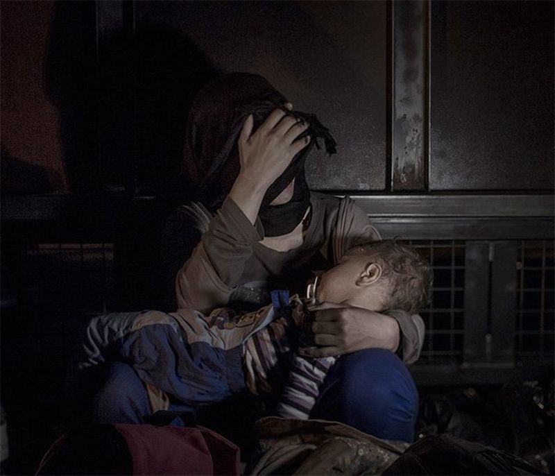 A mother holds her sleeping baby as she sits against a metal gate, covering her face in distress.