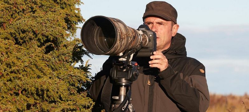 Photographer Markus Varesvuo positions a Canon camera with a long lens, which is on a tripod and covered with a camouflage lens cover. He stands beside a fir tree.