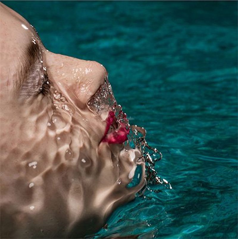 A close-up portraits shows a model with red lipstick emerging from water, light shimmering on the coating of water on her face.