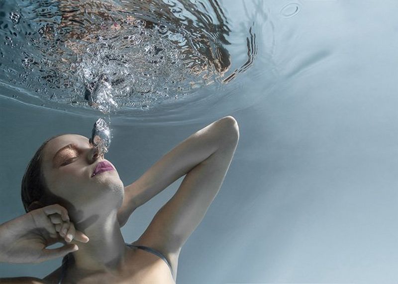 A woman submerged underwater with hands behind her raised head as large bubbles from her mouth break the surface above her.