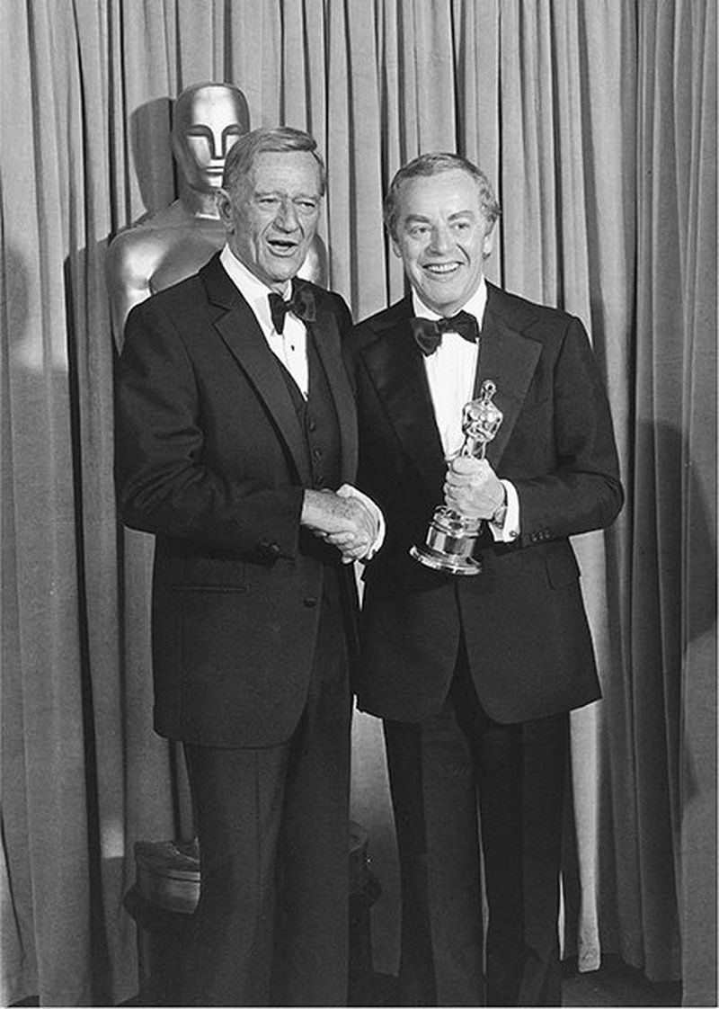 John Wayne and Michael Deeley shaking hands at the Oscars, Michael holding an award.