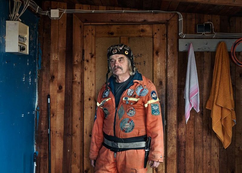 A man in an orange boiler suit stands in a room with a wooden-panelled wall, two towels hanging from pegs.