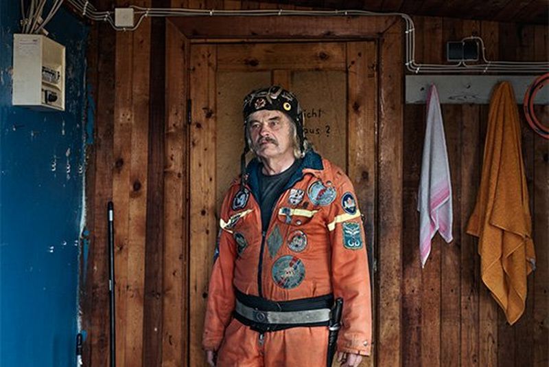 A man in an orange boiler suit stands in a room with a wooden-panelled wall, two towels hanging from pegs.