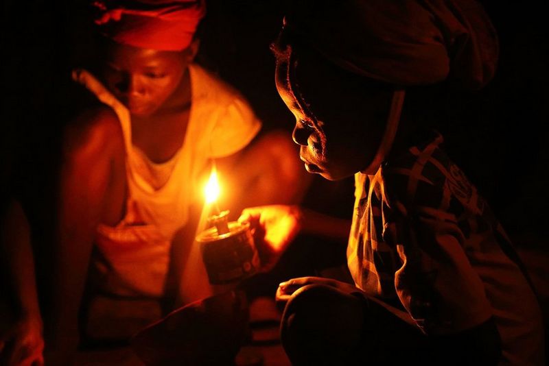 A child crouches and holds an oil lamp in the dark.