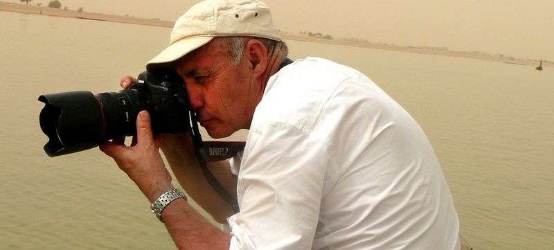 Photographer Pascal Maitre points his Canon camera at a subject we can’t see. He stands in front of the sea.