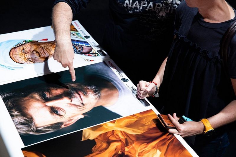People look at three photos on a display - the left, of yellow flame-like flowing fabric, the middle, a close-up of a middle-aged man, the right of an older African woman in colourful printed clothes
