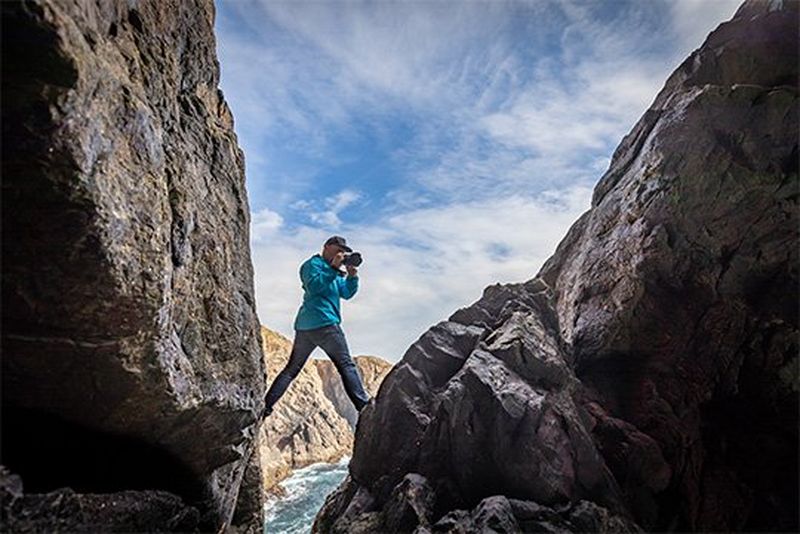 Action photographer Richard Walch shooting with a Canon EOS R, one leg on each side of a steep coastal cliff.