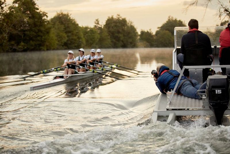 Matthew Joseph, lies on a motorised boat with his camera poised to photograph rowers.