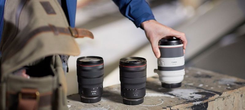 A hand places a Canon RF 70-200mm F2.8L IS USM lens on a plank alongside an RF 24-70mm F2.8L IS USM and RF 15-35mm F2.8L IS USM lens.