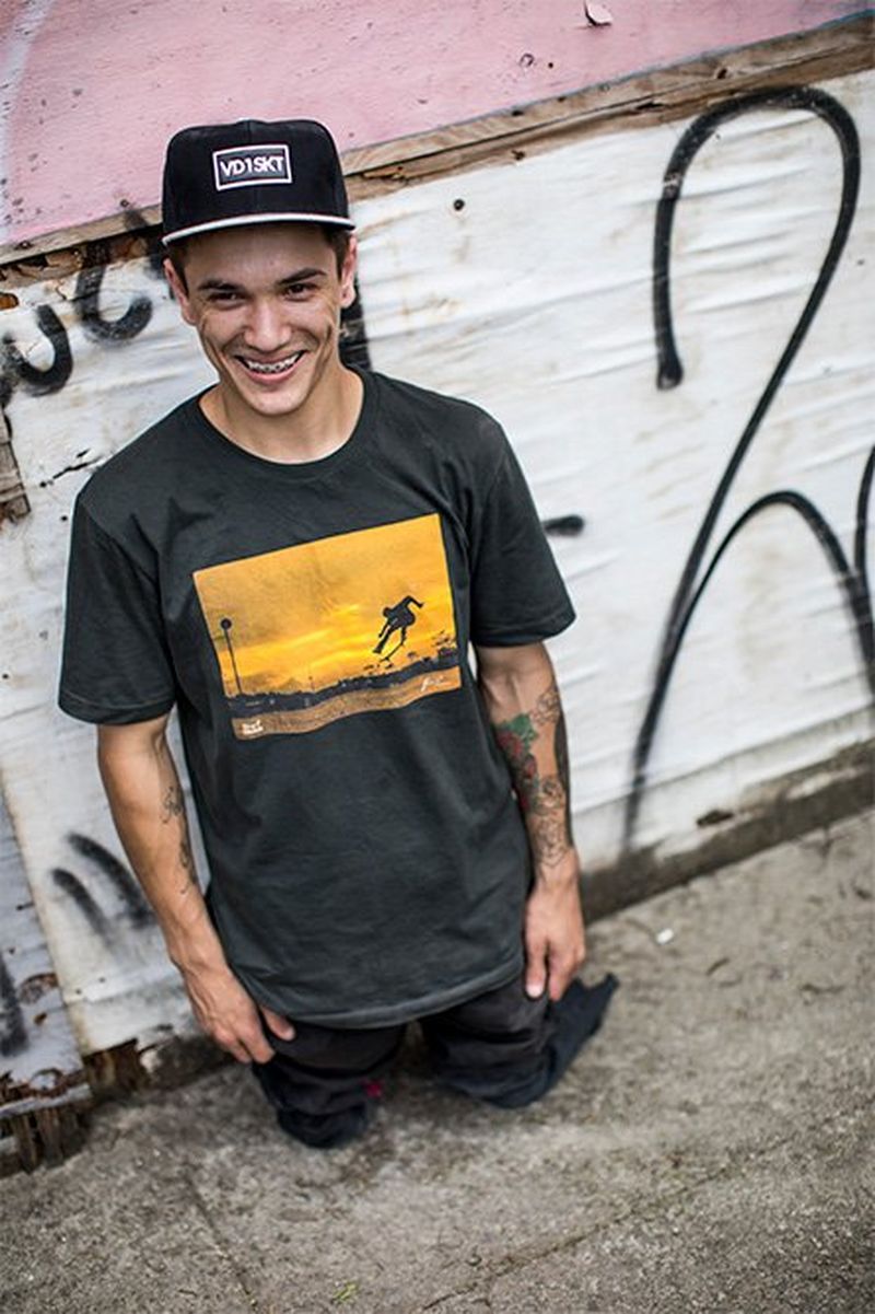 Double amputee Felipe Nunes stands, without prosthetic legs, against a graffiti-covered wall – he's smiling casually, with a cap on. 