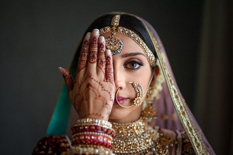 An Indian woman in silk wedding clothes and opulent gold jewellery holds a henna tattooed hand in front of one eye.