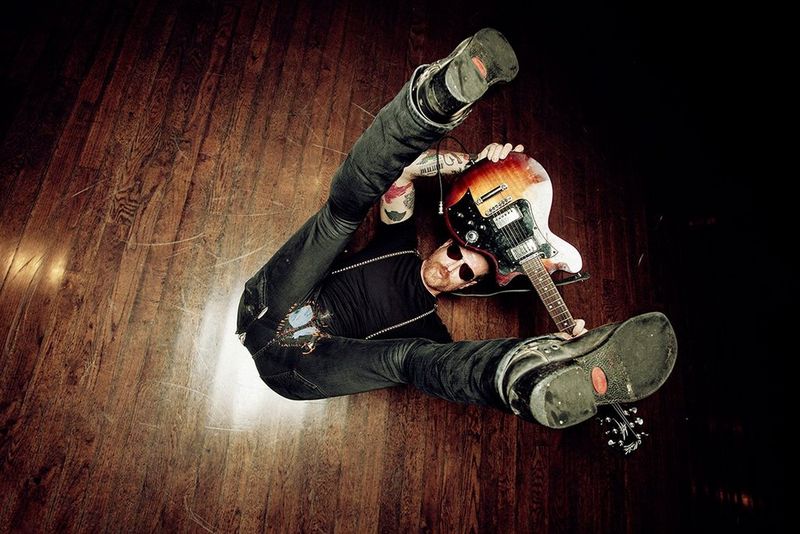 Guitarist Jesse Hughes lying on his back on a wooden floor, legs raised, holding a guitar above his head with both hands