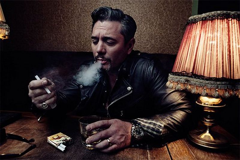 Huey Morgan, wearing a black leather jacket, sits at a wooden table, blowing smoke from a cigarette