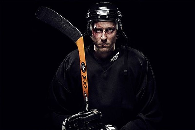 An ice hockey player staring intently, holding his stick in both gloved hands, his face looking cut and bruised