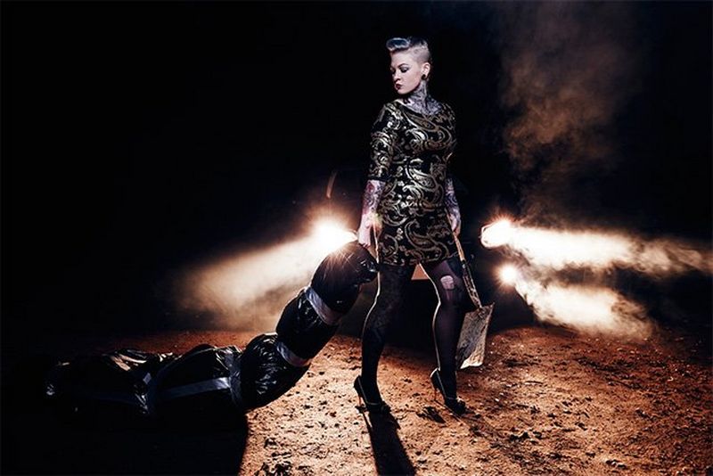 A tattooed woman in black carries a shovel in one hand and drags a body wrapped in black bin bags sealed with tape