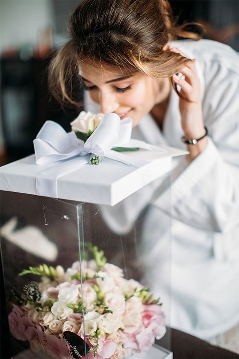 A woman in a white dressing gown bends over to sniff flowers on a box tied with a large bow.