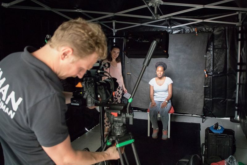 A young woman sits in a black-draped ready to be interviewed on camera.