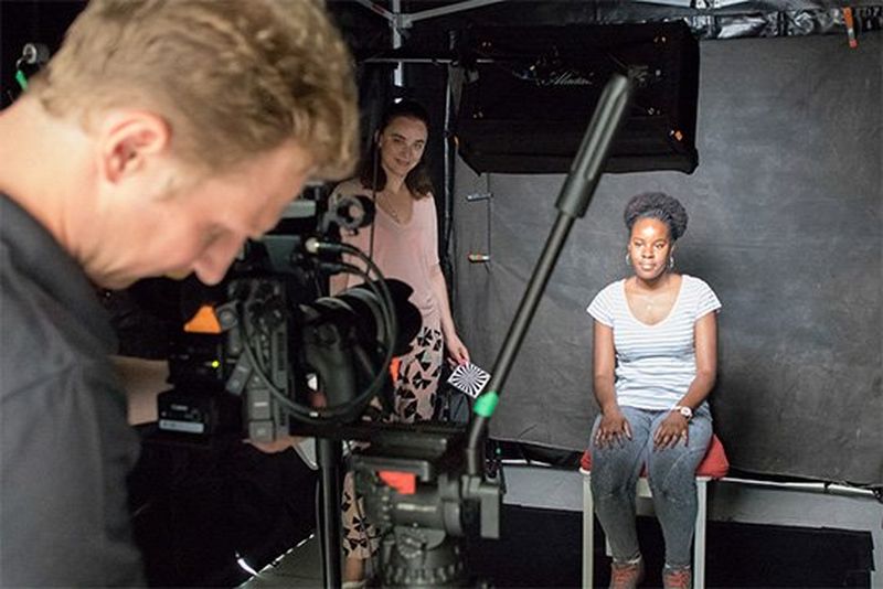 A young woman sits in a black-draped ready to be interviewed on camera.