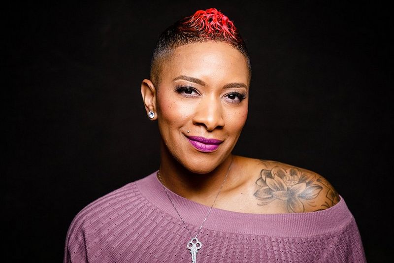 A woman with short, dyed red hair and tattoos wears a lilac top.