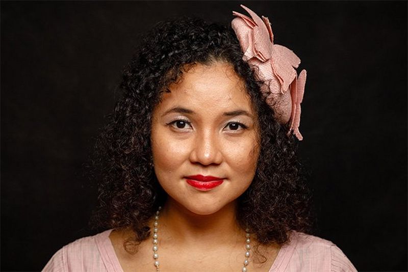 A woman wears a pink top, a large pink flower in her dark, curly hair, and a pearl necklace.