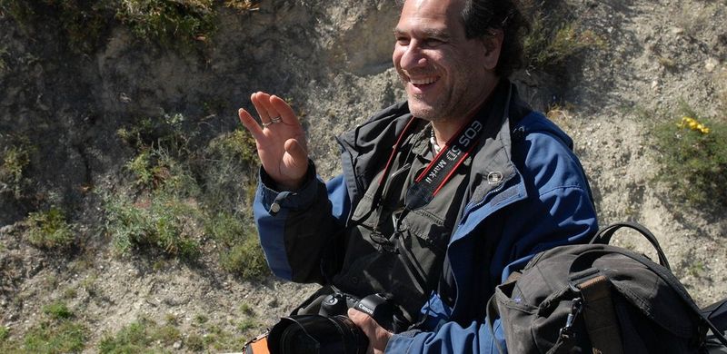 Photographer Patrick Zachmann holds a Canon DSLR and wears a waterproof jacket. He stands in front of some bushes.