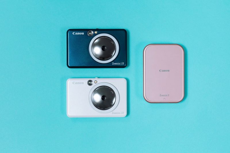 A rose gold Canon Zoemini 2 sits on a bright blue background next to a white and a teal Canon Zoemini S2.