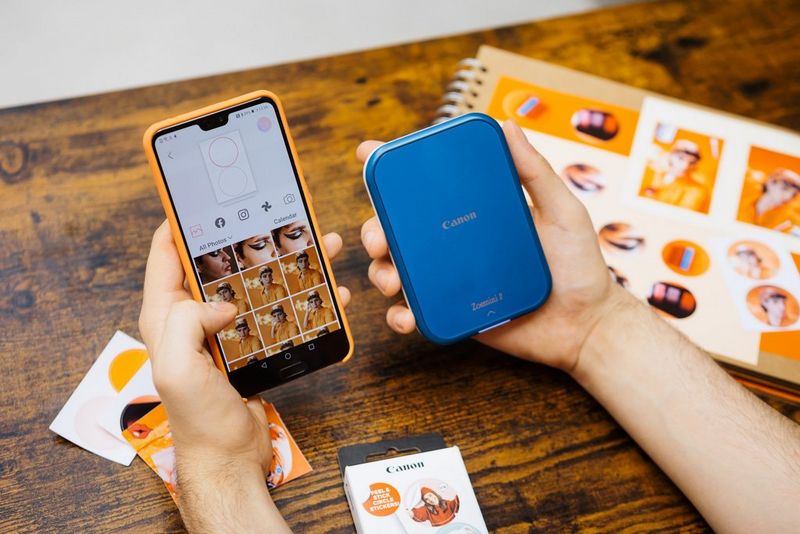 Una persona tiene in una mano uno smartphone che mostra l'app Mini Print e nell'altra una stampante Canon Zoemini 2. Sul tavolo sullo sfondo sono presenti diverse stampe fotografiche.