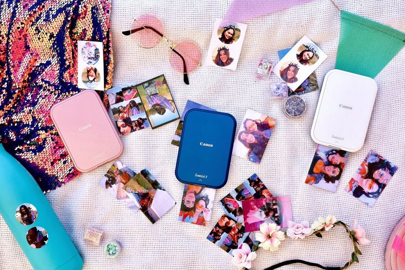 Tri štampača Canon Zoemini 2 stoje usred odštampanih fotografija, naočara za sunce, traka za kosu i boca za vodu na raznobojnoj podlozi.