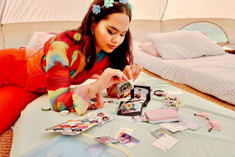 A woman dressed in music festival attire peels off a sticker from a sheet of stickers, while lying on a bed surrounded by more stickers. Next to her a Canon Zoemini 2 portable printer is printing out another sheet of stickers.