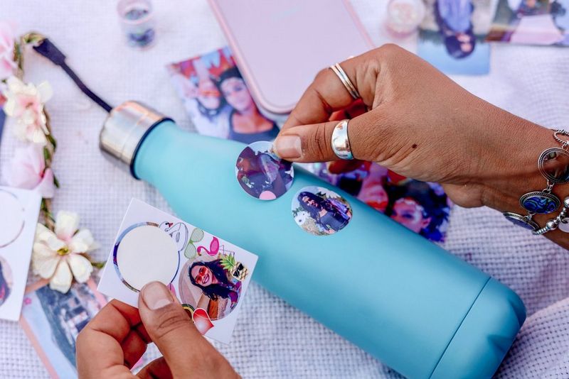 Hands sticking circular photo stickers on a pale blue water bottle, with a Canon Zoemini printer behind.